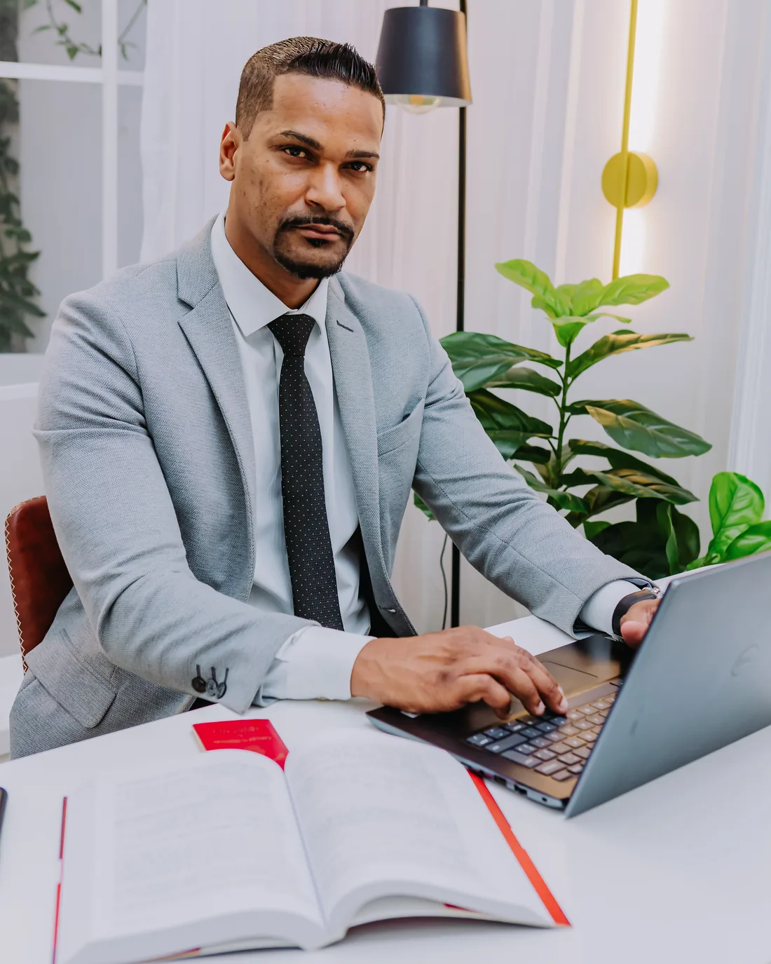 Dr. Marciano Souza à mesa de trabalho, de blazer e gravata, com notebook e um código aberto à frente, olhando diretamente para a câmera